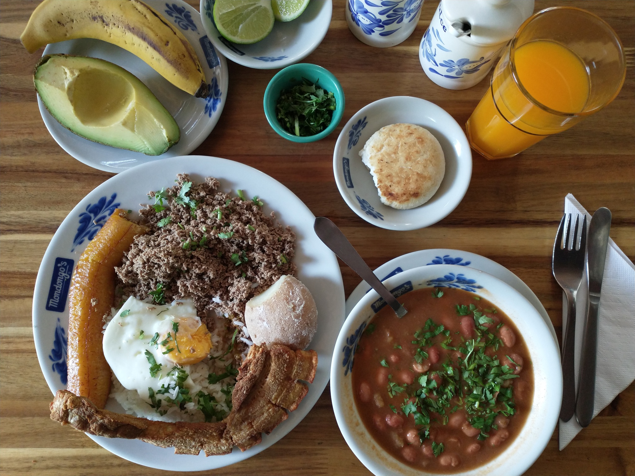 Bandeja paisa lunch platter with avocado, banana, lime, herbs, maize arepa, orange juice, chicharrones, egg, ground beef, potato, and pinto beans.